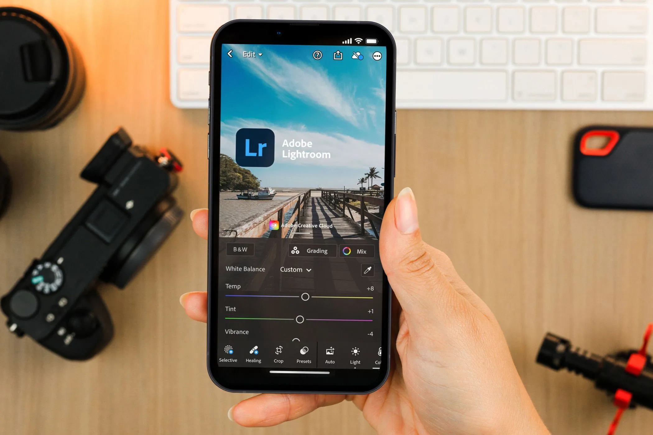 Bàn tay cầm điện thoại hiển thị ứng dụng Lightroom Mobile, phía sau là thiết bị máy ảnh chuyên nghiệp