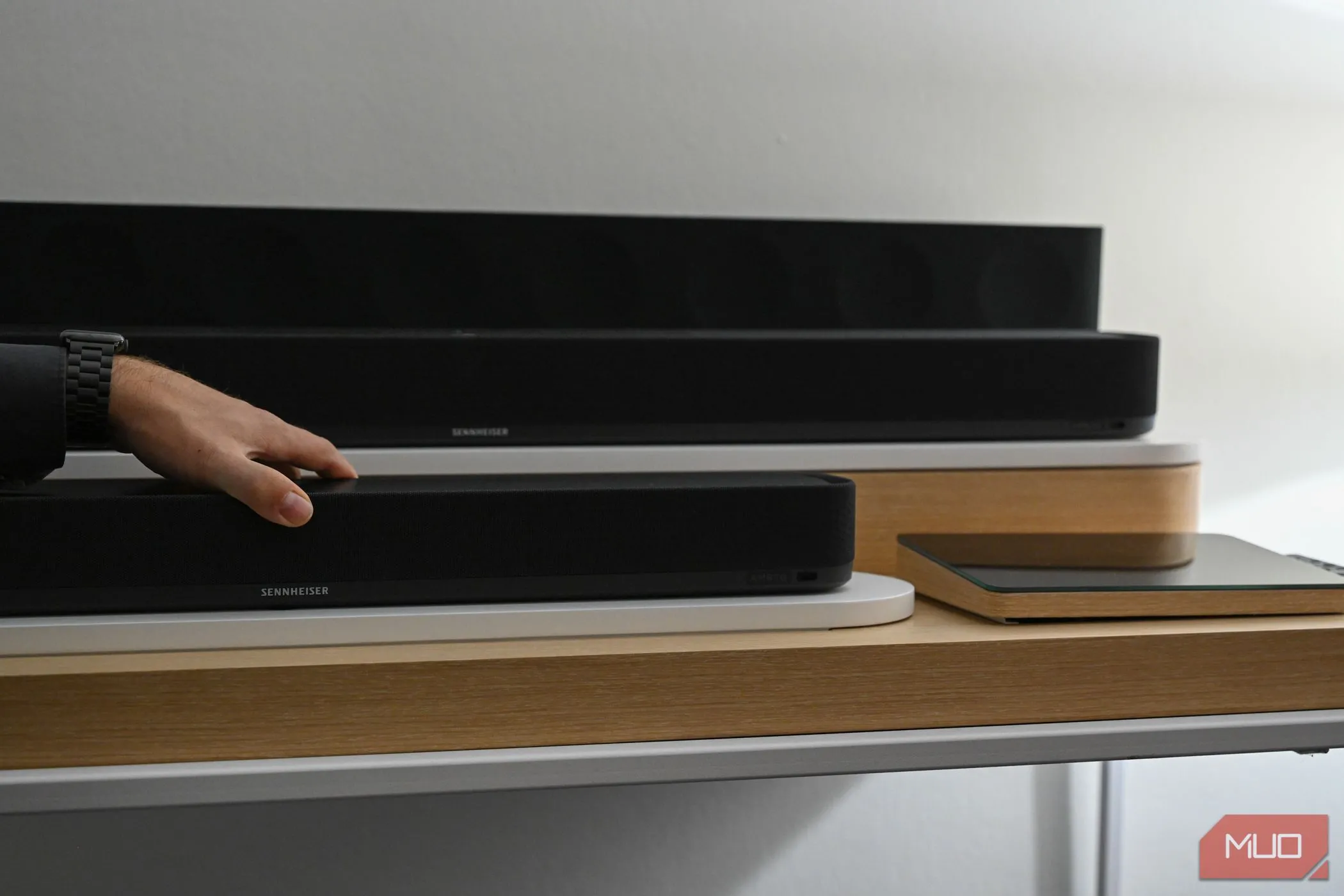 Bàn tay đặt trên loa soundbar Sennheiser Ambeo Soundbar Mini, tượng trưng cho việc người dùng phải thao tác trực tiếp để bật, tắt hoặc điều chỉnh loa.